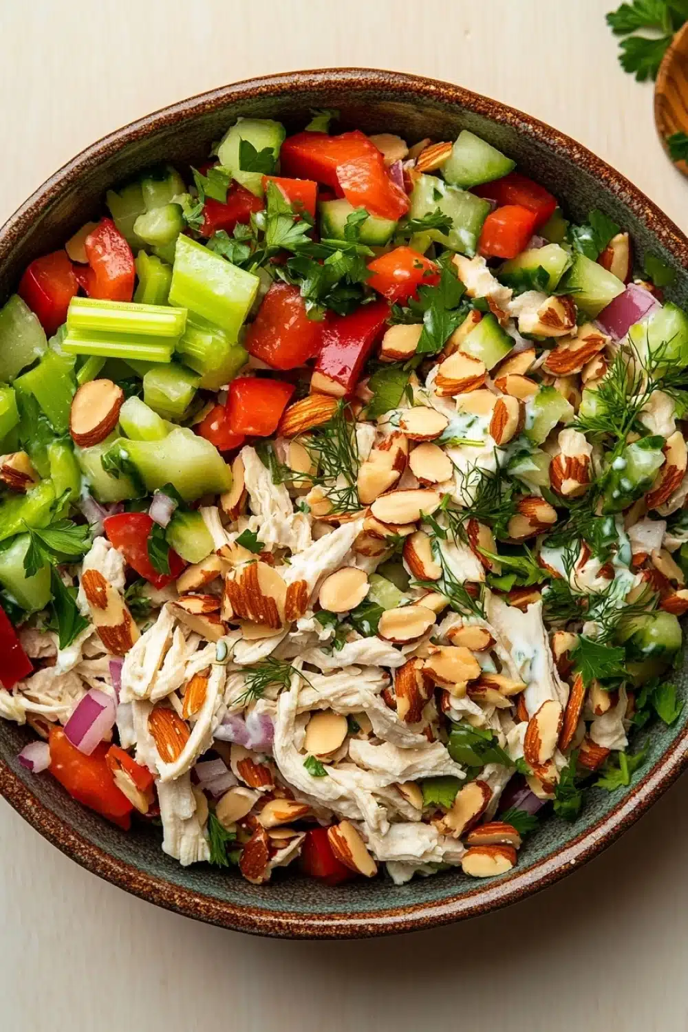 Chicken salad no mayo greek yogurt - the image is of a large bowl filled with a colorful salad. the salad is made up of various ingredients such as sliced cucumbers, red bell peppers, sliced onions, sliced almonds, and dill. the ingredients are mixed together in a creamy dressing, which appears to be chicken or chicken salad. there are also some fresh herbs and spices scattered throughout the salad, adding a pop of green color to the dish. the bowl is sitting on a wooden table, and there is a sprig of parsley on the side.