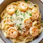 Creamy cajun shrimp scampi over pasta - the image is a close-up of a bowl of pasta dish. the pasta appears to be fettuccine alfredo, with chunks of shrimp scattered on top. the dish is garnished with chopped parsley and grated parmesan cheese. the bowl is made of ceramic and is sitting on a wooden table. the background is blurred, but it seems to be a kitchen countertop.