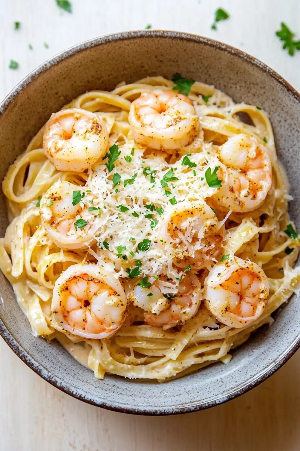 Creamy cajun shrimp scampi over pasta - the image is a close-up of a bowl of pasta dish. the pasta appears to be fettuccine alfredo, with chunks of shrimp scattered on top. the dish is garnished with chopped parsley and grated parmesan cheese. the bowl is made of ceramic and is sitting on a wooden table. the background is blurred, but it seems to be a kitchen countertop.