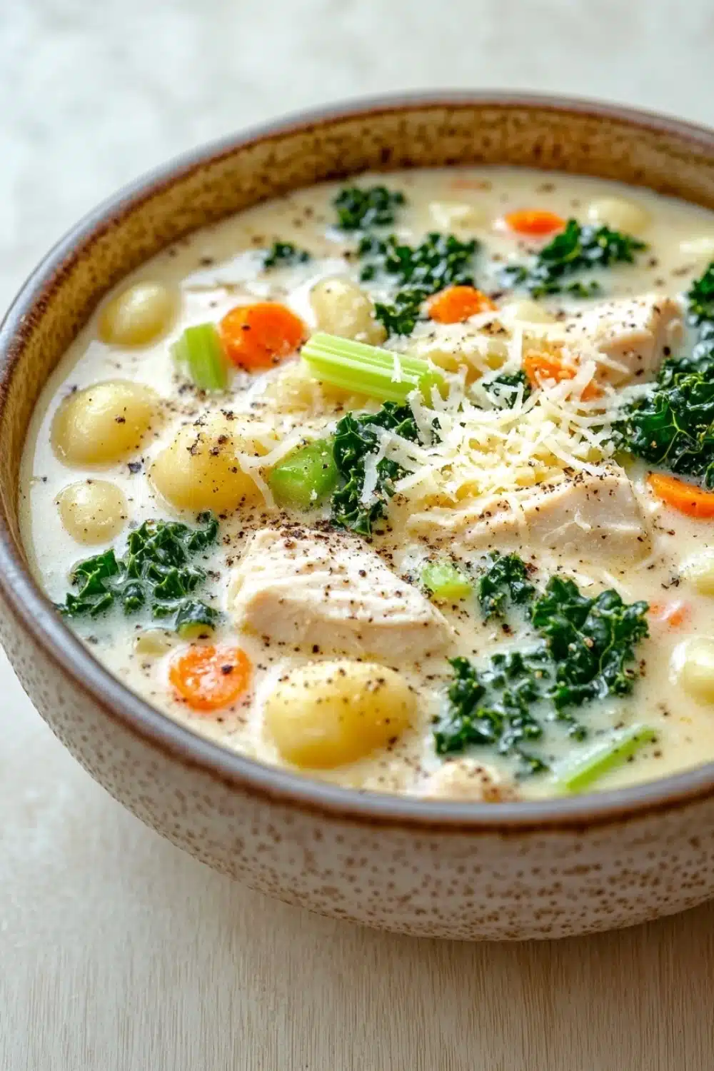 Creamy chicken gnocchi kale soup - the image is a close-up of a bowl of soup. the soup appears to be a creamy white color with chunks of chicken and vegetables mixed in. the bowl is made of ceramic and has a speckled pattern on the rim. there are also small chunks of carrots, celery, and kale scattered throughout the soup. on top of the soup, there is a sprinkle of grated cheese. the background is a light beige color, and the bowl is sitting on a wooden surface.