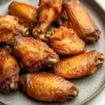 Crispy chicken wings baked - the image shows a plate of baked chicken wings. the wings are golden brown and appear to be crispy on the outside. they are arranged in a circular pattern on the plate, with some overlapping each other. the plate is made of a light-colored ceramic or porcelain material with a speckled texture. the background is a white marble countertop.