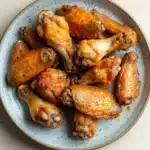 Crispy chicken wings baked - the image shows a plate of cooked chicken wings. the wings are golden brown and appear to be seasoned with herbs and spices. they are arranged in a circular pattern on the plate, with some overlapping each other. the plate is white with a light blue speckled pattern around the edges. the background is a wooden table.