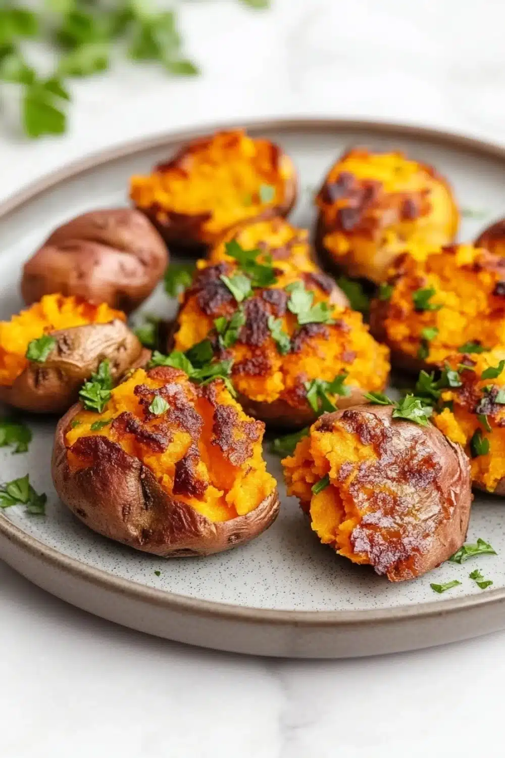 Crispy smashed sweet potatoes air fryer - the image shows a plate of roasted sweet potatoes. the potatoes are golden brown and have a crispy texture. they are garnished with chopped parsley, adding a pop of green color to the dish. the plate is white and the background is blurred, making the potatoes the focal point of the image.