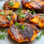 Crispy smashed sweet potatoes air fryer - the image shows a plate of roasted sweet potatoes. the sweet potatoes are golden brown and appear to be seasoned with herbs and spices. they are arranged in a circular pattern on the plate, with some overlapping each other. the plate is made of light-colored wood and has a speckled texture. there are also a few sprigs of fresh parsley scattered around the plate. the background is blurred, but it appears to be a wooden table.