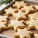 Crispy sugar cookie cut out - the image shows a baking tray lined with freshly baked star-shaped cookies. the cookies are golden brown in color and have a light dusting of powdered sugar on top. the tray is placed on a wooden table with a small christmas tree in the background. the tree is decorated with small white lights, giving it a warm and cozy feel. the overall mood of the image is festive and cozy.