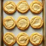 Crispy sugar cookie cut out - the image shows a baking tray lined with parchment paper. on the tray, there are nine round cookies arranged in a single row. the cookies appear to be freshly baked and have a golden brown color. each cookie has a crumbly texture on top, with a small indentation in the center. the parchment paper is white and has a textured surface. the tray is sitting on a white marble countertop.