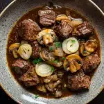 Crock pot beef mushrooms - the image is a close-up of a bowl of beef stew. the beef is cooked medium-rare and appears to be tender and juicy. it is garnished with sliced onions and mushrooms. the stew is a rich brown color and looks thick and flavorful. the bowl is made of ceramic and is sitting on a dark wooden table. the overall appearance of the dish is hearty and appetizing.