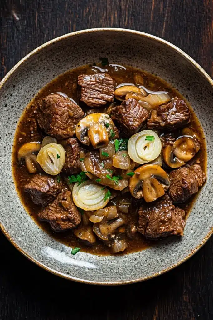 Crock pot beef mushrooms - the image is a close-up of a bowl of beef stew. the beef is cooked medium-rare and appears to be tender and juicy. it is garnished with sliced onions and mushrooms. the stew is a rich brown color and looks thick and flavorful. the bowl is made of ceramic and is sitting on a dark wooden table. the overall appearance of the dish is hearty and appetizing.
