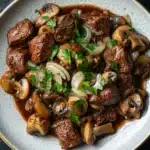 Crock pot beef mushrooms - the image is a close-up of a plate of food. the plate is white with a gold rim and is placed on a black table. the food appears to be a dish made with chunks of beef, sliced onions, and mushrooms. the beef is cooked in a rich, dark brown sauce and is garnished with chopped parsley. the mushrooms are sliced and have a light brown color. the dish looks appetizing and ready to eat.