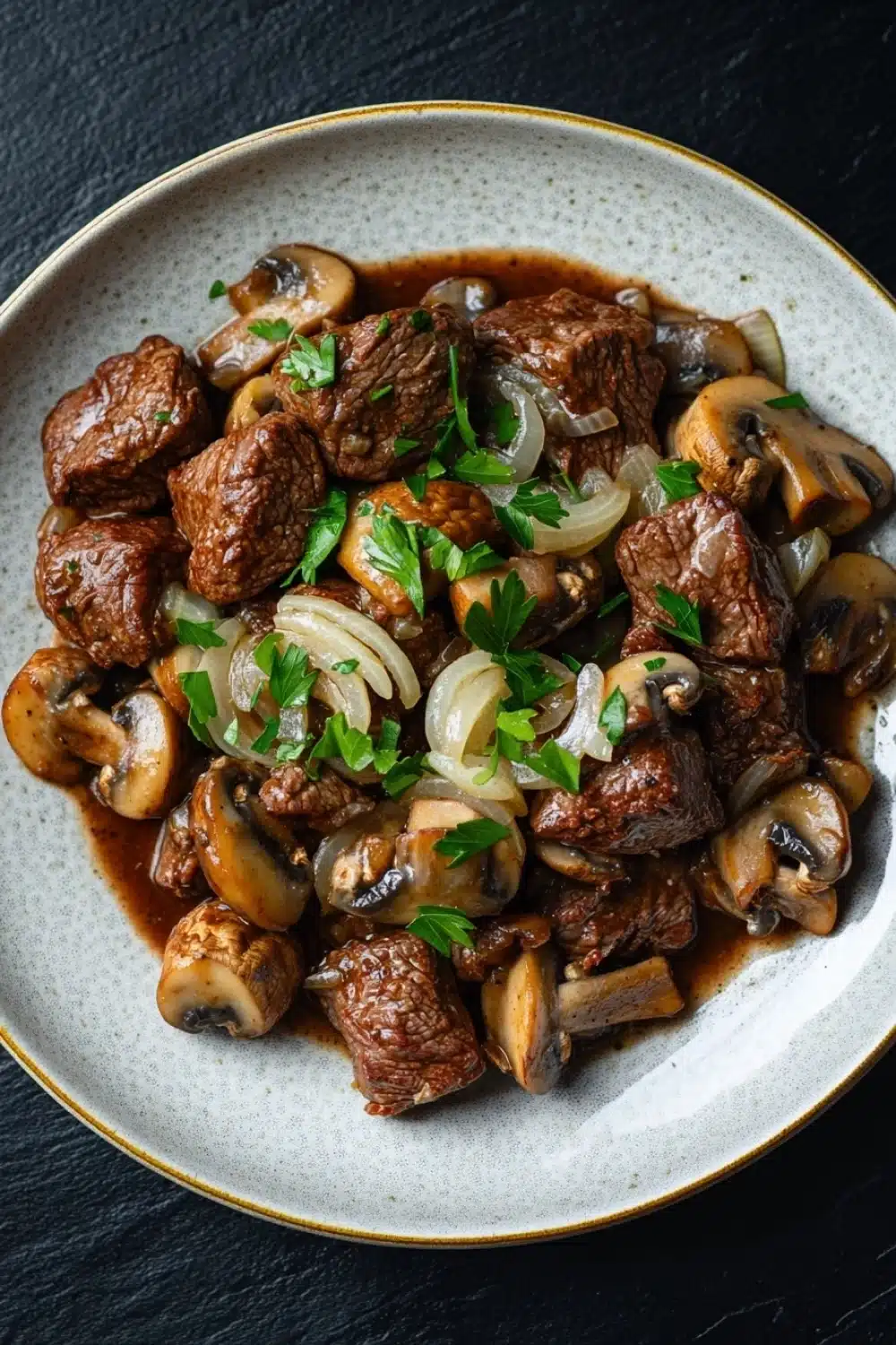 Crock pot beef mushrooms - the image is a close-up of a plate of food. the plate is white with a gold rim and is placed on a black table. the food appears to be a dish made with chunks of beef, sliced onions, and mushrooms. the beef is cooked in a rich, dark brown sauce and is garnished with chopped parsley. the mushrooms are sliced and have a light brown color. the dish looks appetizing and ready to eat.