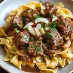Crock pot beef over noodles - the image shows a plate of beef stroganoff with fettuccine noodles. the beef is cooked in a rich, dark brown sauce and is garnished with chopped parsley. the noodles are long and thin, and they are arranged in a pile on top of each other. the dish is served on a dark grey plate with a textured rim. the background is blurred, but it appears to be a kitchen countertop.