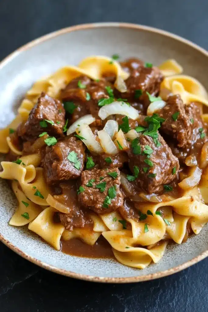 Crock pot beef over noodles - the image shows a plate of beef stroganoff with fettuccine noodles. the beef is cooked in a rich, dark brown sauce and is garnished with chopped parsley. the noodles are long and thin, and they are arranged in a pile on top of each other. the dish is served on a dark grey plate with a textured rim. the background is blurred, but it appears to be a kitchen countertop.