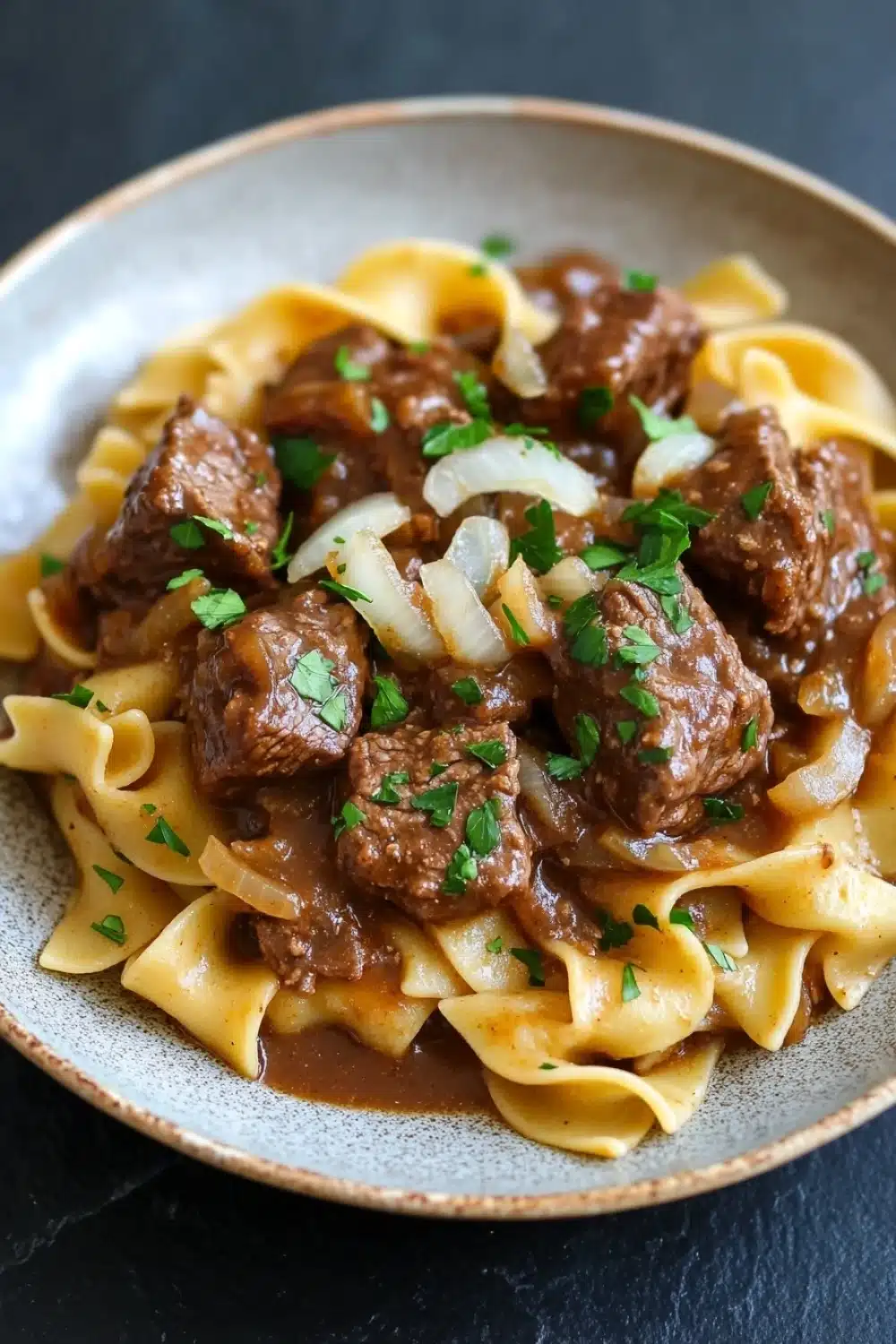 Crock pot beef over noodles - the image shows a plate of beef stroganoff with fettuccine noodles. the beef is cooked in a rich, dark brown sauce and is garnished with chopped parsley. the noodles are long and thin, and they are arranged in a pile on top of each other. the dish is served on a dark grey plate with a textured rim. the background is blurred, but it appears to be a kitchen countertop.