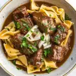Crock pot beef over noodles - the image is a close-up of a bowl of beef stroganoff. the beef is cooked in a rich, dark brown sauce and is garnished with chopped parsley and sliced onions. the noodles are long and thin, and they are arranged in a pile on top of the beef. the bowl is made of ceramic and has a light brown color. the background is a white marble countertop.