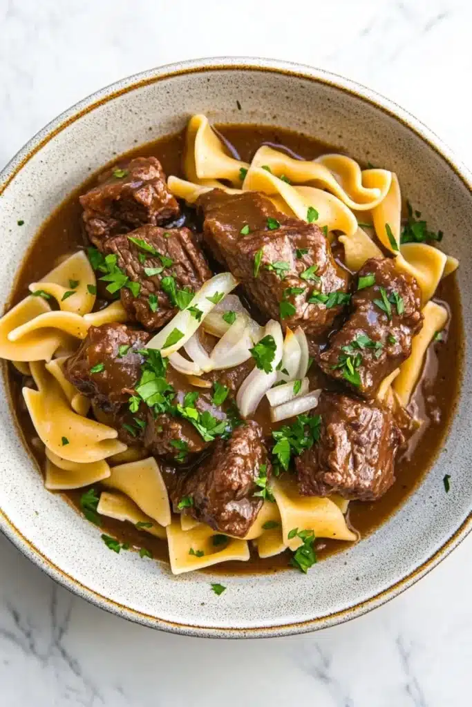 Crock pot beef over noodles - the image is a close-up of a bowl of beef stroganoff. the beef is cooked in a rich, dark brown sauce and is garnished with chopped parsley and sliced onions. the noodles are long and thin, and they are arranged in a pile on top of the beef. the bowl is made of ceramic and has a light brown color. the background is a white marble countertop.
