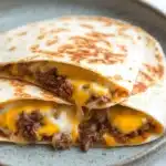 Crock pot beef quesadilla - the image shows two quesadillas stacked on top of each other on a gray plate. the quesads are made with a tortilla shell and are filled with ground beef, cheese, and other ingredients. the cheese is melted and oozing out of the sides of the tortillas. the plate is sitting on a white marble countertop.