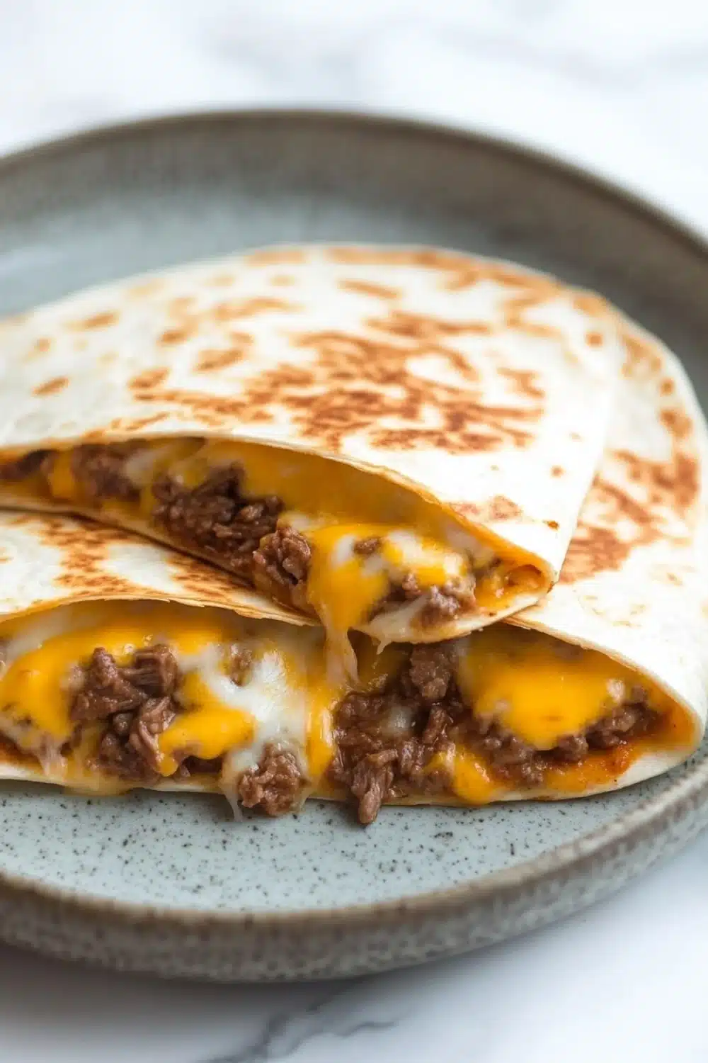 Crock pot beef quesadilla - the image shows two quesadillas stacked on top of each other on a gray plate. the quesads are made with a tortilla shell and are filled with ground beef, cheese, and other ingredients. the cheese is melted and oozing out of the sides of the tortillas. the plate is sitting on a white marble countertop.