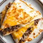 Crock pot beef quesadilla - the image shows a plate with two quesadillas stacked on top of each other. the plate is white with a blue rim and has a speckled pattern around the edges. the quesads are golden brown and appear to be freshly made. they are filled with shredded beef, onions, and cheese. the cheese is melted and oozing out of the sides of the plate. the background is a white marble countertop.