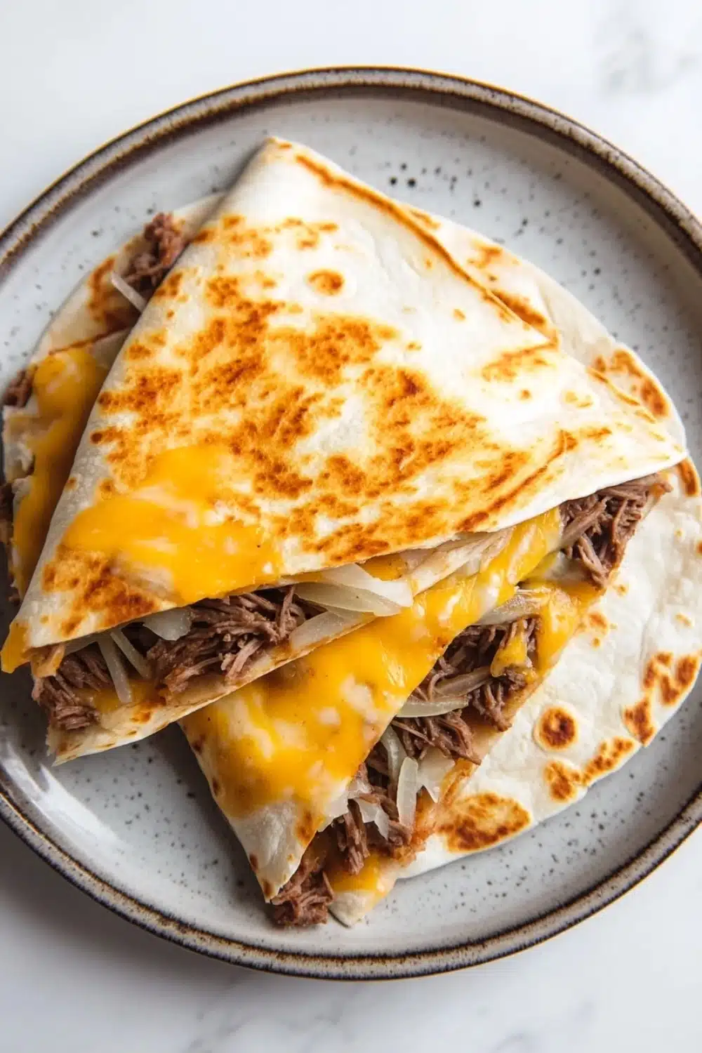 Crock pot beef quesadilla - the image shows a plate with two quesadillas stacked on top of each other. the plate is white with a blue rim and has a speckled pattern around the edges. the quesads are golden brown and appear to be freshly made. they are filled with shredded beef, onions, and cheese. the cheese is melted and oozing out of the sides of the plate. the background is a white marble countertop.