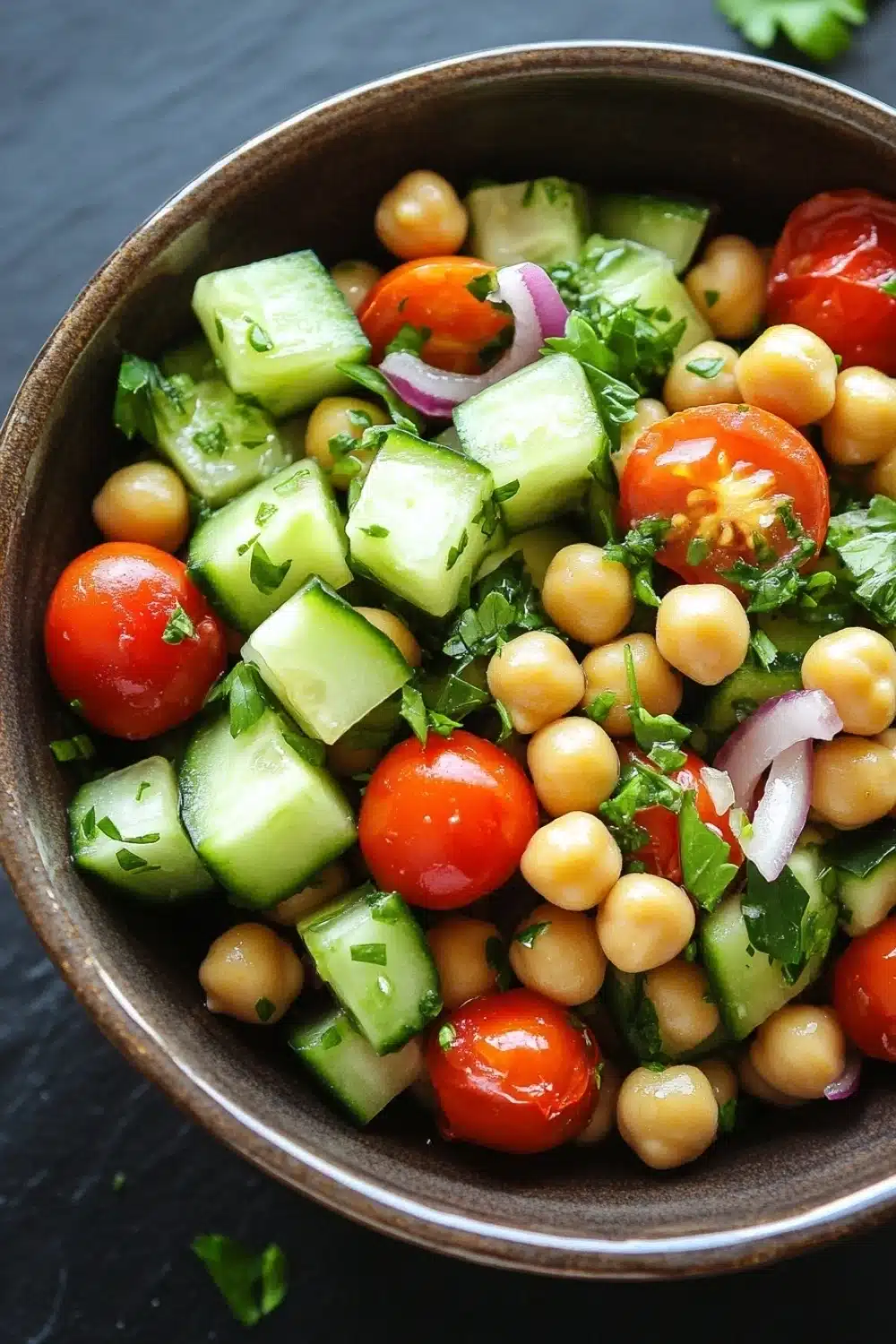 Cucumber tomato salad garbanzo - the image is a close-up of a bowl of salad. the bowl is made of dark brown ceramic and is placed on a dark grey countertop. the salad is made up of various ingredients, including sliced cucumbers, cherry tomatoes, chickpeas, and red onions. there are also some fresh cilantro leaves scattered throughout the salad.