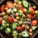 Cucumber tomato salad garbanzo - the image is a close-up of a bowl of salad. the bowl is made of dark brown ceramic and is placed on a wooden table. the salad is made up of diced tomatoes, cucumbers, chickpeas, red onions, and fresh herbs. the tomatoes are bright red and appear to be ripe and juicy. the cucumbers are cut into small cubes and are scattered throughout the salad, while the red onions are sliced into thin strips. the herbs are finely chopped and add a pop of green color to the dish. there are also a few sprigs of parsley scattered around the bowl.