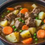 Freezer beef stew crock pot - the image is a close-up of a bowl of beef stew. the stew is made with chunks of beef, potatoes, carrots, and peas. the beef appears to be cooked medium-rare and has a reddish-brown color. the potatoes are yellow and appear to be seasoned with herbs and spices. the carrots are bright orange and the peas are green. the bowl is made of dark brown ceramic and is sitting on a wooden table. the background is blurred, making the stew the focal point of the image.