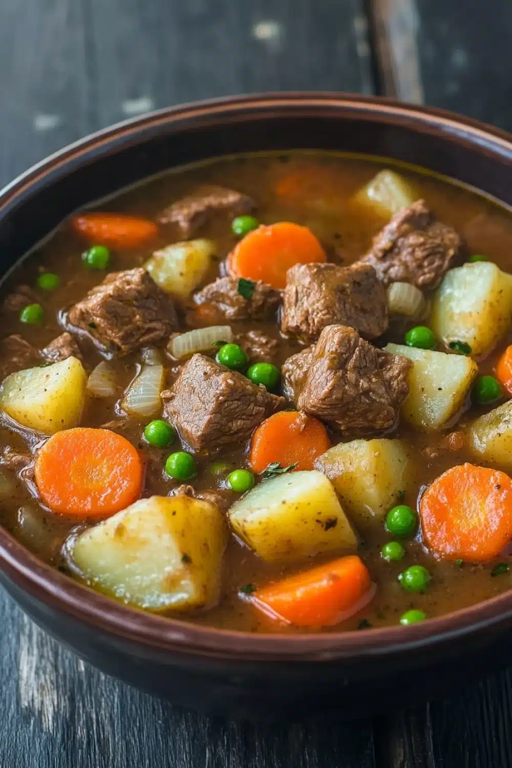 Freezer beef stew crock pot - the image is a close-up of a bowl of beef stew. the stew is made with chunks of beef, potatoes, carrots, and peas. the beef appears to be cooked medium-rare and has a reddish-brown color. the potatoes are yellow and appear to be seasoned with herbs and spices. the carrots are bright orange and the peas are green. the bowl is made of dark brown ceramic and is sitting on a wooden table. the background is blurred, making the stew the focal point of the image.
