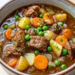 Freezer beef stew crock pot - the image is a close-up of a bowl of beef stew. the stew is made with chunks of beef, potatoes, carrots, peas, and onions. the beef is cooked in a rich, dark brown broth and is garnished with chopped herbs. the vegetables are bright orange and appear to be fresh. the bowl is made of ceramic and is sitting on a wooden table. the background is blurred, so the focus is on the stew.