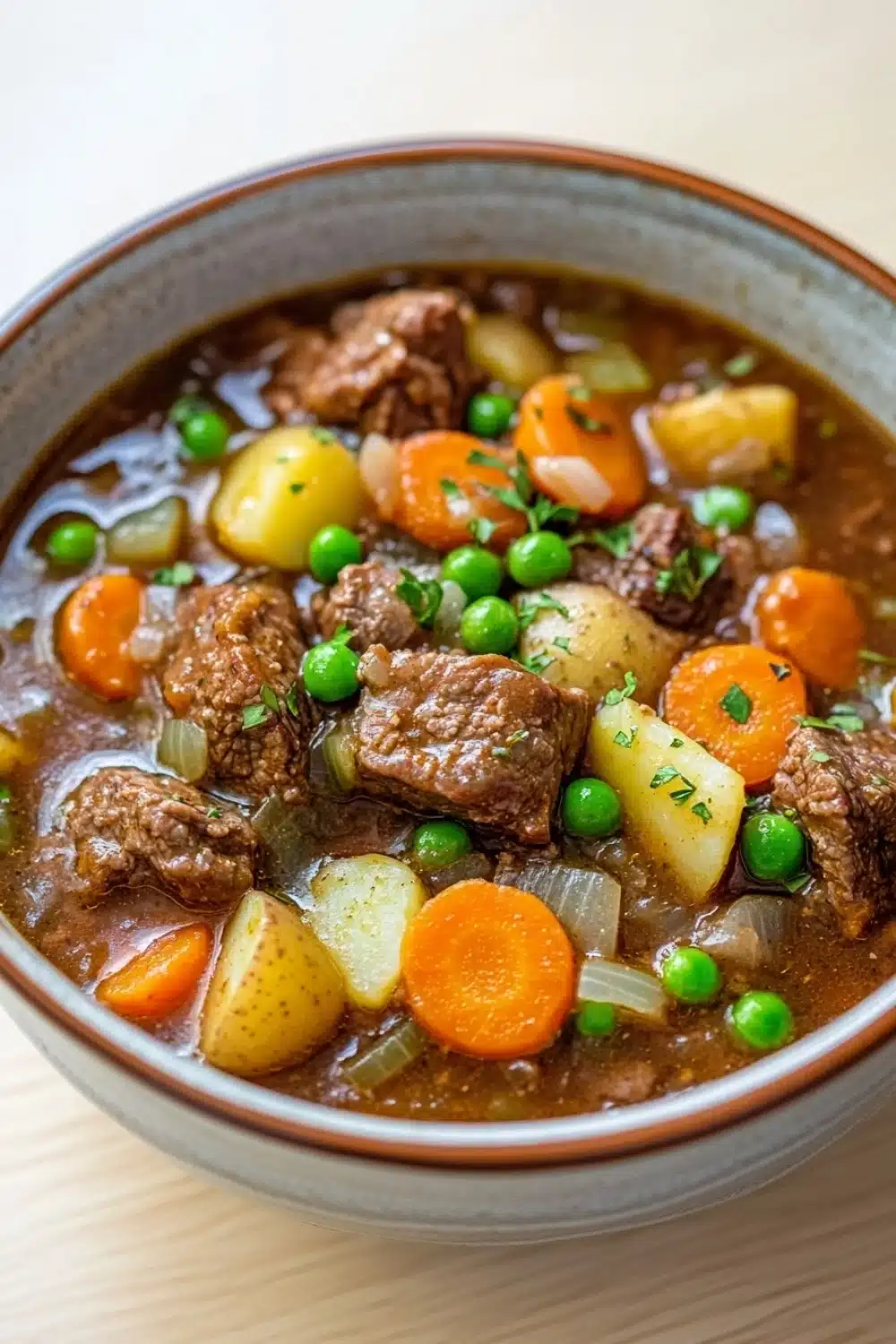 Freezer beef stew crock pot - the image is a close-up of a bowl of beef stew. the stew is made with chunks of beef, potatoes, carrots, peas, and onions. the beef is cooked in a rich, dark brown broth and is garnished with chopped herbs. the vegetables are bright orange and appear to be fresh. the bowl is made of ceramic and is sitting on a wooden table. the background is blurred, so the focus is on the stew.