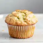 Green apple muffin - the image is of a freshly baked muffin on a white plate. the muffin is golden brown in color and has a crumbly texture. it appears to be a muffin with a light dusting of powdered sugar on top. the plate is sitting on a marble countertop with a gray background. the overall mood of the image is warm and inviting.