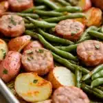 Green bean sausage sheet pan dinner - the image shows a baking tray filled with cooked potatoes and green beans. the potatoes are golden brown and appear to be seasoned with herbs and spices. the green beans are long and slender, and they are arranged in a pile on top of the potatoes. there are also some small pieces of sausage scattered throughout the dish. the tray is lined with parchment paper, and the background is blurred, but it appears to be a kitchen countertop.