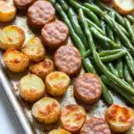 Green bean sausage sheet pan dinner - the image shows a baking tray filled with roasted potatoes and green beans. the potatoes are golden brown and appear to be seasoned with herbs and spices. the green beans are long and slender, and they are arranged in a pile on the tray. the tray is lined with parchment paper, and the background is white. the overall appearance of the dish is savory and appetizing.