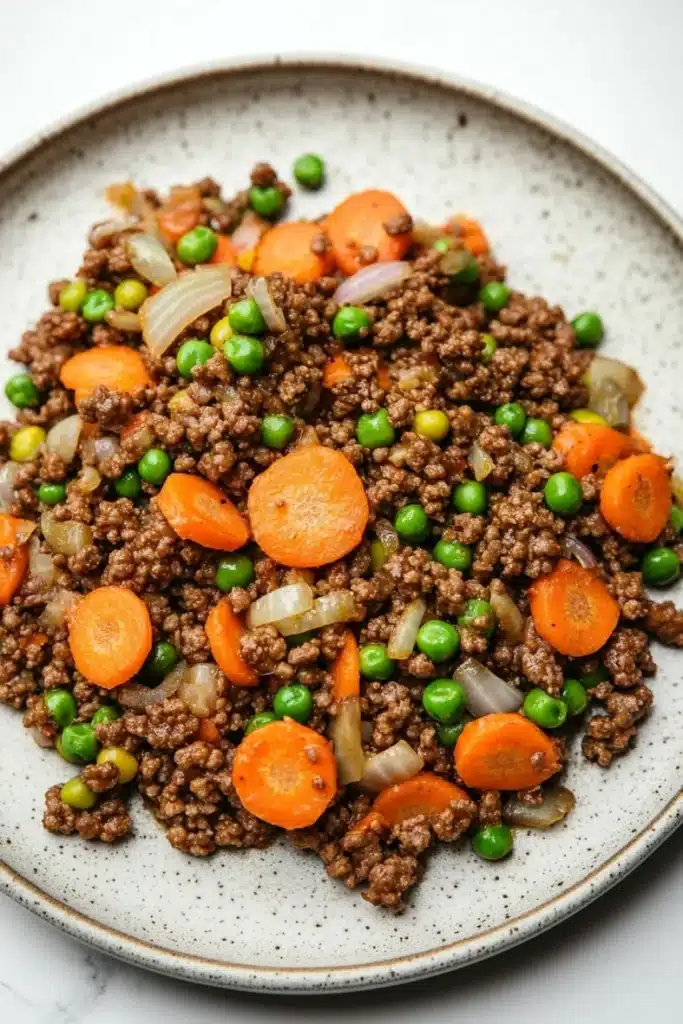 Ground beef dinner for kids - the image is a close-up of a plate of food. the plate is round and has a light brown color with a speckled texture. on top of the plate, there is a mixture of ground beef, carrots, peas, and onions. the beef appears to be cooked and is mixed with the vegetables. the carrots are bright orange and the peas are green. the onions are white and are scattered throughout the dish. the dish looks fresh and appetizing.