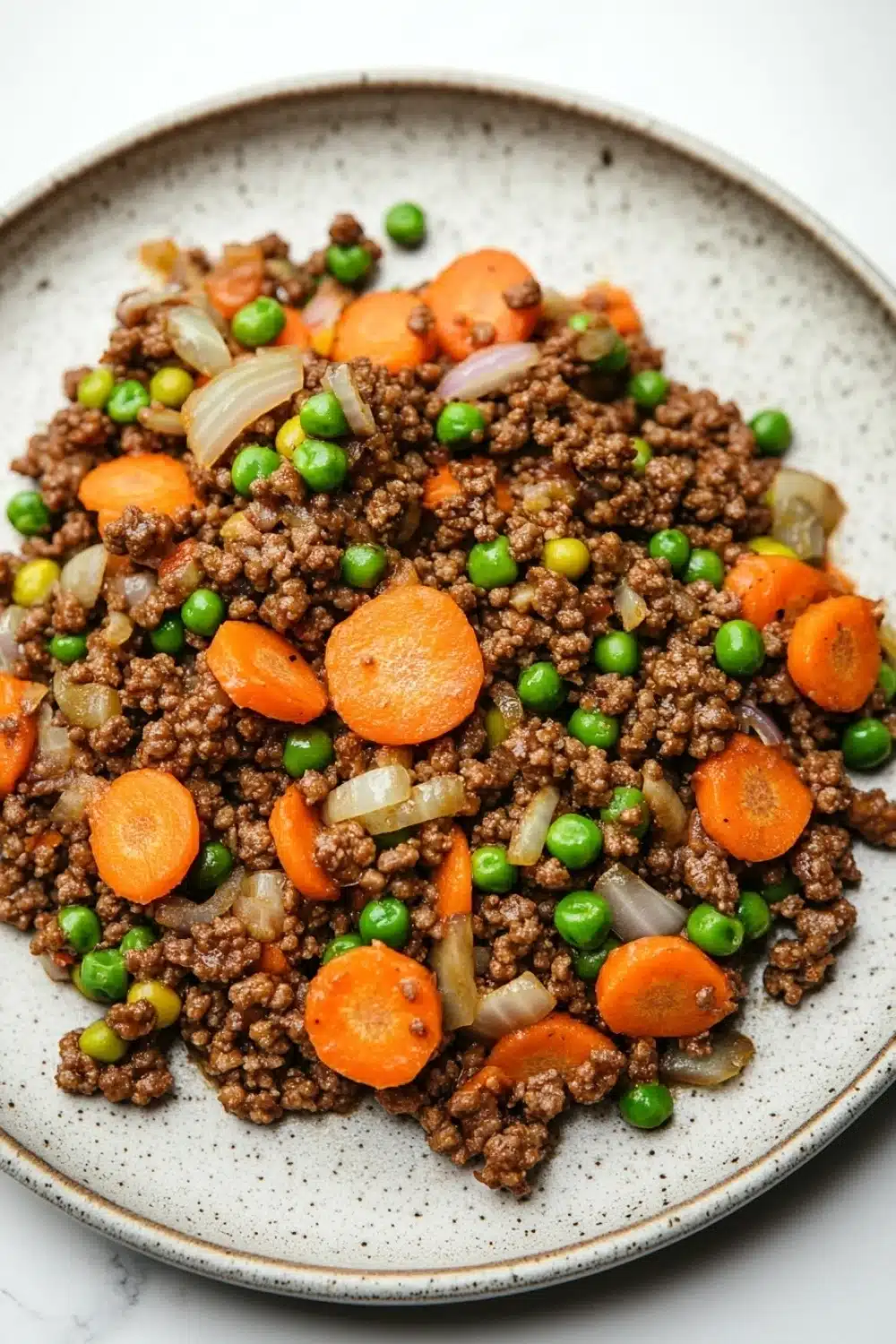Ground beef dinner for kids - the image is a close-up of a plate of food. the plate is round and has a light brown color with a speckled texture. on top of the plate, there is a mixture of ground beef, carrots, peas, and onions. the beef appears to be cooked and is mixed with the vegetables. the carrots are bright orange and the peas are green. the onions are white and are scattered throughout the dish. the dish looks fresh and appetizing.