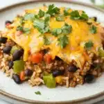 High protein beef enchilada skillet - the image shows a plate of enchiladas on a white marble countertop. the enchillas are covered in a layer of melted cheese and are garnished with fresh cilantro. the cheese is golden brown and appears to be melted and bubbly. on top of the cheese, there are chunks of ground beef, black beans, red bell peppers, and green bell peppers. the dish is served on a bed of brown rice. the plate is white with a speckled pattern and there are a few sprigs of cilantro scattered around the plate.