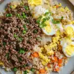 High protein korean beef fried rice - the image is a close-up of a plate of food. the plate is made of a light-colored ceramic material and is placed on a wooden table. the food appears to be a type of rice dish with a variety of ingredients. on top of the rice, there are chunks of ground beef, diced carrots, and sliced onions. there are also two hard-boiled eggs on top, which are yellow and white in color. the dish is garnished with chopped green onions and sesame seeds. the overall appearance of the dish is colorful and appetizing.