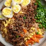 High protein korean beef fried rice - the image is a close-up of a plate of food. the plate is round and has a light brown color. on top of the plate, there is a bed of brown rice with chunks of ground beef, sliced carrots, and green peas scattered throughout. there are also two hard-boiled eggs on top. the dish appears to be a stir-fry or stir fry. the background is a wooden table.