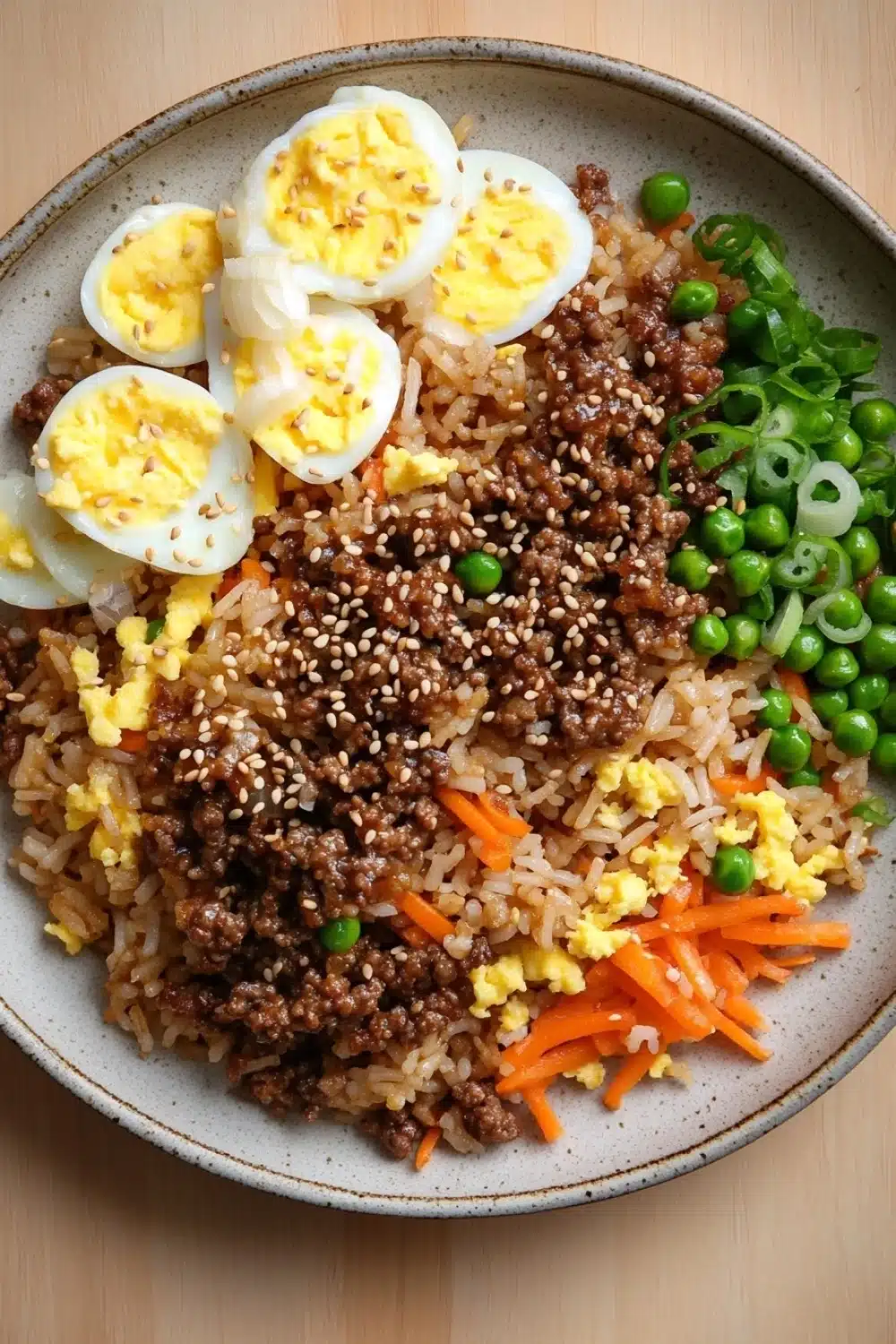 High protein korean beef fried rice - the image is a close-up of a plate of food. the plate is round and has a light brown color. on top of the plate, there is a bed of brown rice with chunks of ground beef, sliced carrots, and green peas scattered throughout. there are also two hard-boiled eggs on top. the dish appears to be a stir-fry or stir fry. the background is a wooden table.