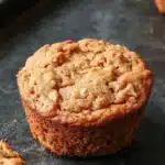 High protein vegan breakfast muffin - the image shows a freshly baked muffin on a black baking tray. the muffin is round and golden brown in color, with a crumbly texture on top. it appears to be freshly baked and has a soft, fluffy interior. the tray is lined with parchment paper, and there are a few more muffins visible in the background. the overall mood of the image is warm and inviting.