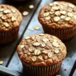 High protein vegan breakfast muffin - the image shows a tray of freshly baked muffins on a wooden table. the muffins are golden brown in color and have a crumbly texture. they are topped with chopped oats, giving them a crunchy appearance. the tray is lined with parchment paper and there are scattered oats around the tray. the background is blurred, but it appears to be a kitchen countertop.