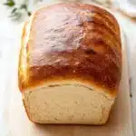 Homemade sandwich bread - the image shows a freshly baked loaf of bread on a wooden cutting board. the bread is golden brown in color and has a soft, fluffy texture. it is placed on a white wooden table with a white cloth napkin and a sprig of fresh thyme on the side. the background is blurred, but it appears to be a kitchen countertop.