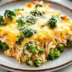 Keto chicken broccoli casserole - the image shows a plate of food with a casserole dish on it. the dish appears to be made with chicken, broccoli, and cheese. the chicken is covered in melted cheese and there are small pieces of broccoli scattered on top. the plate is white with a brown rim and is sitting on a white countertop. in the background, there is a bottle of wine and a bunch of fresh broccoli.