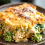 Keto chicken broccoli casserole - the image shows a slice of broccoli casserole on a brown plate. the dish is golden brown and has a layer of melted cheese on top. the cheese appears to be melted and bubbly, and there are small pieces of broccoli scattered around the plate. in the background, there are more broccoli florets, suggesting that the dish is ready to be eaten. the plate is sitting on a wooden table with a blurred background.