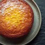 Lemon cake oil - the image is a close-up of a round cake on a gray plate. the cake appears to be freshly baked and has a golden brown crust on top. it is covered in a light dusting of powdered sugar, which is drizzled over the top of the cake. there are small pieces of orange zest scattered across the cake, adding a pop of color to the dish. the plate is placed on a dark grey textured surface.