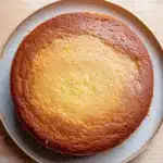 Lemon cake oil - the image is a close-up of a round cake on a light blue plate. the cake appears to be freshly baked and has a golden brown crust on top. the surface of the cake is smooth and shiny, with small yellow specks scattered across it. the plate is placed on a wooden table, and the background is blurred, making the cake the focal point of the image.