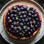 Lemon cake with blueberry pie filling - the image is of a round blueberry tart on a white plate with a speckled pattern. the tart has a golden brown crust and is filled with fresh blueberries. the blueberries are arranged in a circular pattern on top of the tart, with some of them overlapping each other. the plate is placed on a dark wooden table, with a few lemons and green leaves scattered around it. the overall color scheme of the image is dark and earthy, with the blueberries standing out against the white plate.