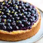 Lemon cake with blueberry pie filling - the image is of a round blueberry cheesecake on a white plate. the cheesecake has a golden brown crust and is topped with a generous amount of fresh blueberries. the blueberries are arranged in a circular pattern on top of the cheesecake, with some of them overlapping each other. the plate is sitting on a wooden table, and the background is blurred, making the blueberries the focal point of the image.
