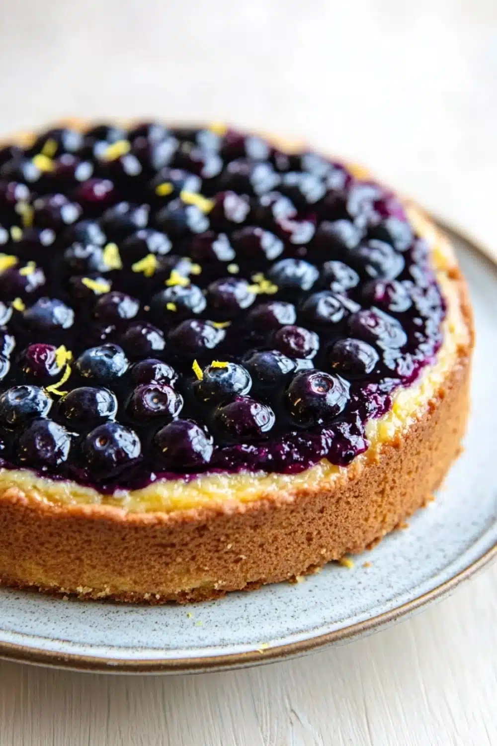 Lemon cake with blueberry pie filling - the image is of a round blueberry cheesecake on a white plate. the cheesecake has a golden brown crust and is topped with a generous amount of fresh blueberries. the blueberries are arranged in a circular pattern on top of the cheesecake, with some of them overlapping each other. the plate is sitting on a wooden table, and the background is blurred, making the blueberries the focal point of the image.