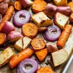 Maple roasted root vegetables - the image shows a baking tray filled with roasted vegetables. the vegetables are cut into small cubes and are arranged in a single layer on the tray. there are also slices of orange and red onions scattered throughout the tray, adding a pop of color to the dish. the tray appears to be made of stainless steel and is sitting on a wooden table. the background is blurred, but it seems to be a kitchen countertop.
