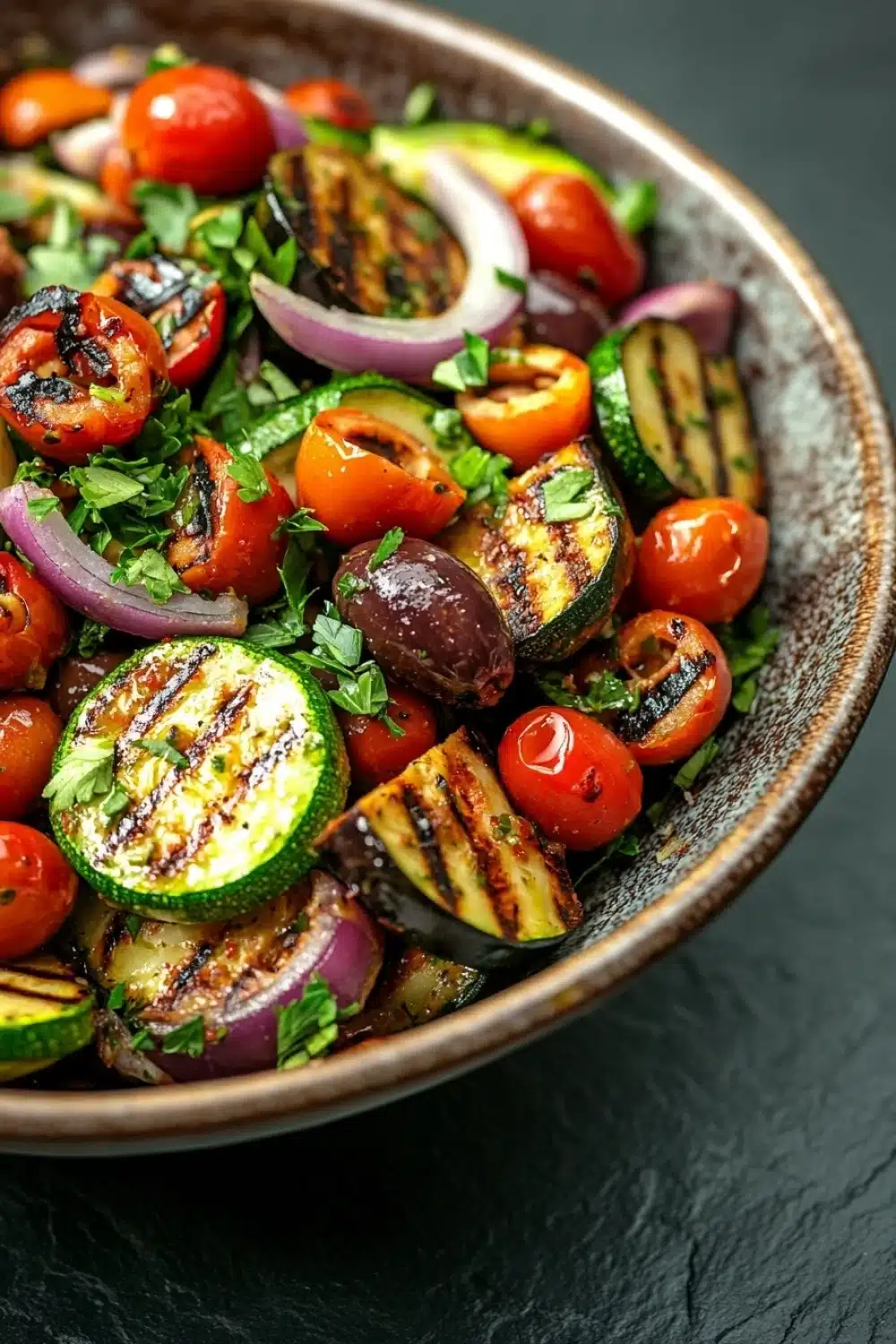 Mediterranean marinated vegetable salad - the image is a close-up of a colorful salad in a bowl. the salad is made up of grilled vegetables, including zucchini, cherry tomatoes, red onions, and olives. the vegetables are arranged in a circular pattern, with some overlapping each other. the colors of the vegetables are vibrant and the texture appears to be fresh and appetizing. the bowl is made of ceramic and has a dark brown finish. the background is a dark grey countertop.